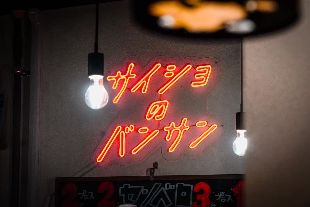 裸電球とともに壁面を照らすサイショのバンサンの赤いLEDネオンサイン